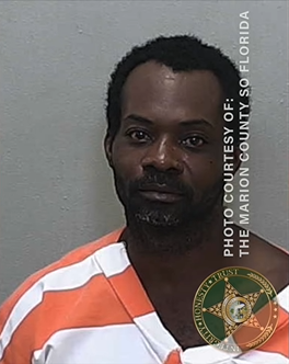 Mugshot of a African American man with short, black hair and a black and grey beard in a orange and white striped jumpsuit against a neutral background.