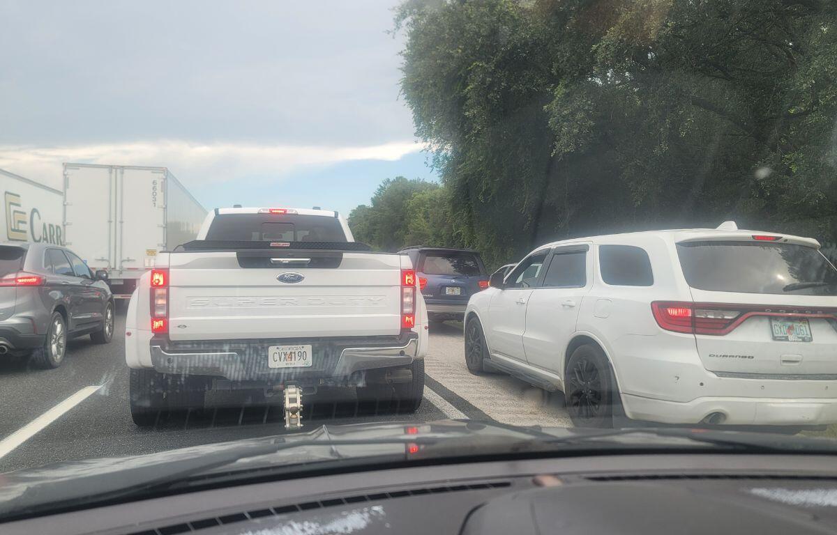 Foreground looking through the windshield at cars to the left, a pick-up truck in the middle front and vehicle to the right. A tree line is to the right.