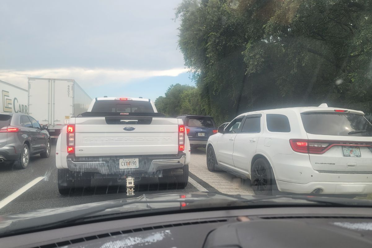 Foreground looking through the windshield at cars to the left, a pick-up truck in the middle front and vehicle to the right. A tree line is to the right.