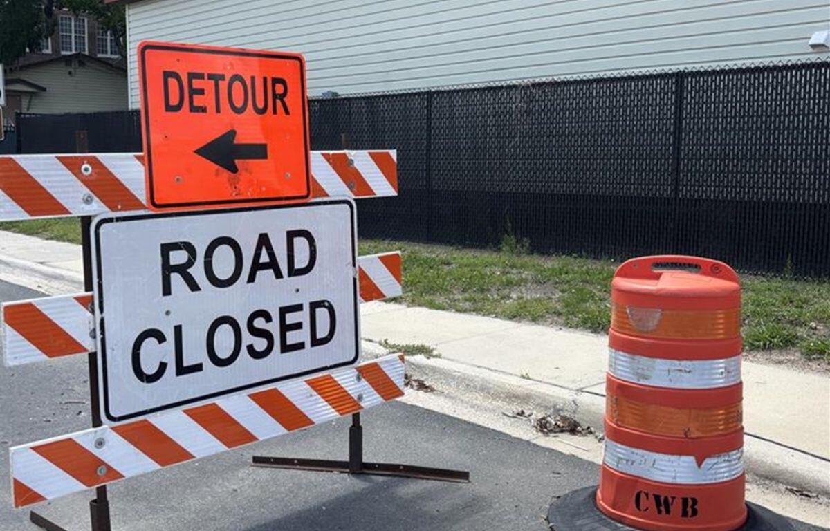 Street signs, orange on top over a white sign, on an orange and white sawhorse, with an orange and white pylon to the right.