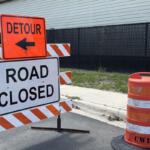 Street signs, orange on top over a white sign, on an orange and white sawhorse, with an orange and white pylon to the right.