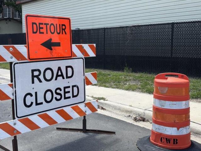 Street signs, orange on top over a white sign, on an orange and white sawhorse, with an orange and white pylon to the right.