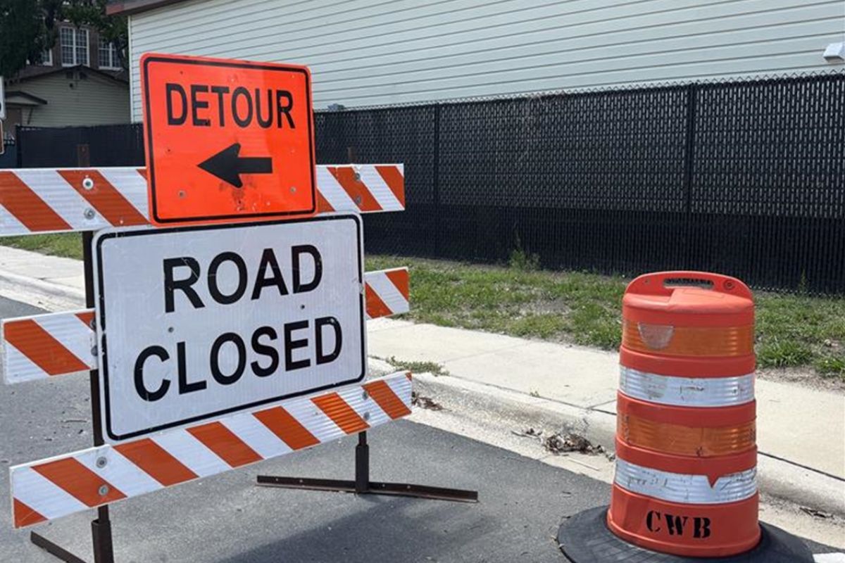 Street signs, orange on top over a white sign, on an orange and white sawhorse, with an orange and white pylon to the right.