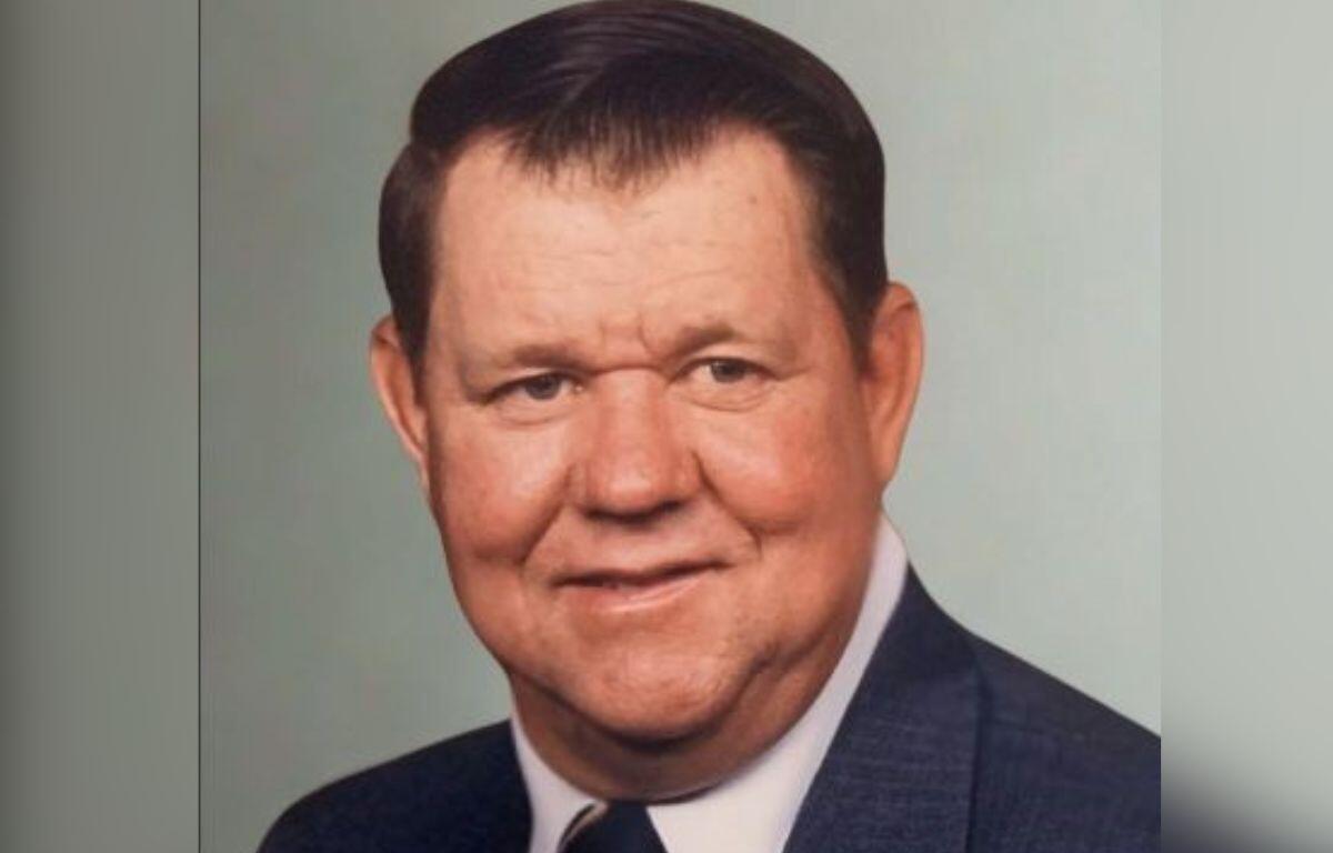 A picture of a man in a suit, with a gray wall int he background.
