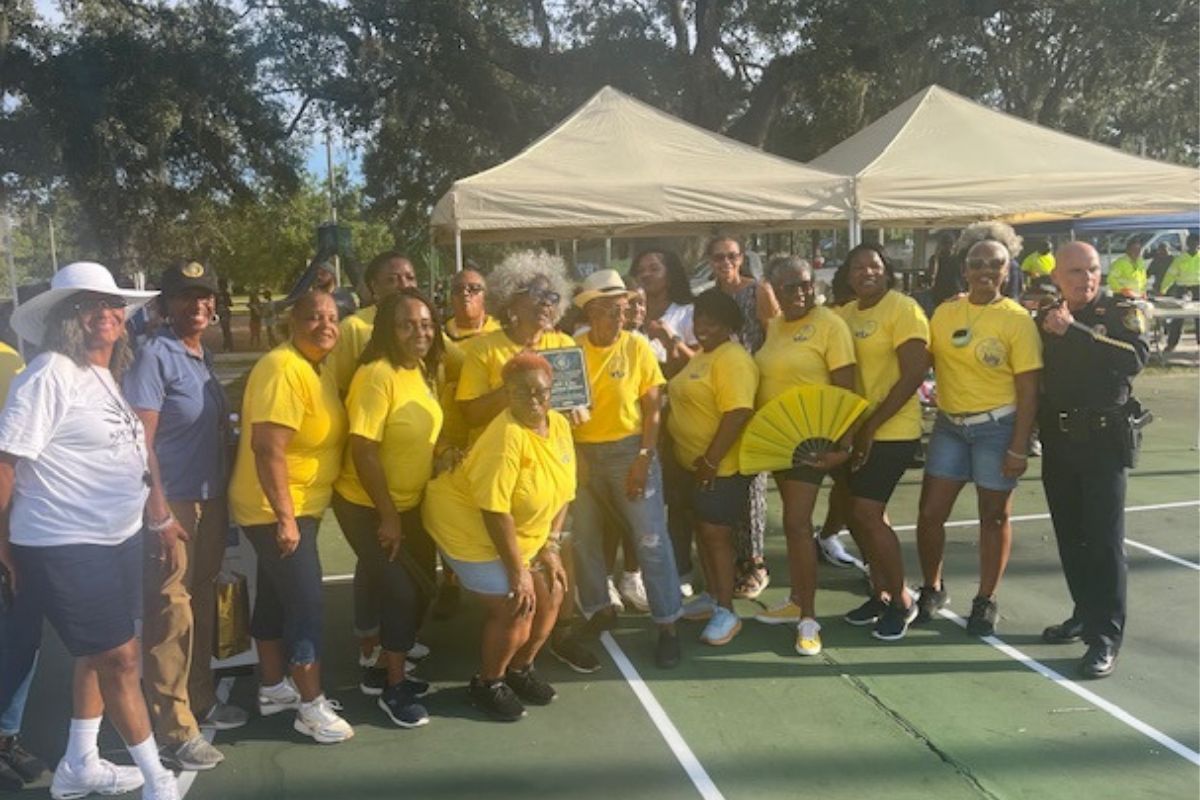 A woman with a white hat and shirt on the extreme left, next to a woman with a baseball hat and blue shirt and women in yellow, next to a policeman, with tents in the background