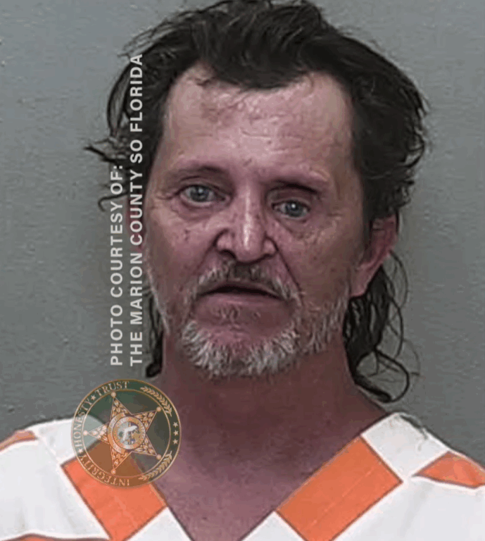 Mugshot of a man with medium-length dark hair wearing an orange-and-white jail uniform, facing forward against a neutral background.