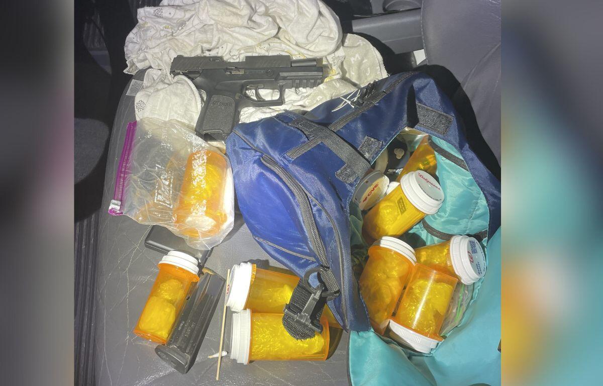 A sidearm on a white piece of cloth, with orange pill containers, plastic baggies, magazines to the sidearm, on top of a blue bag, and gray car seat.