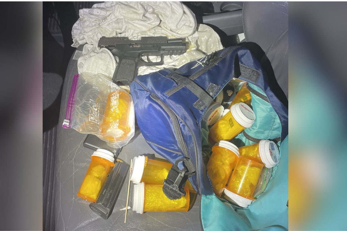 A sidearm on a white piece of cloth, with orange pill containers, plastic baggies, magazines to the sidearm, on top of a blue bag, and gray car seat.