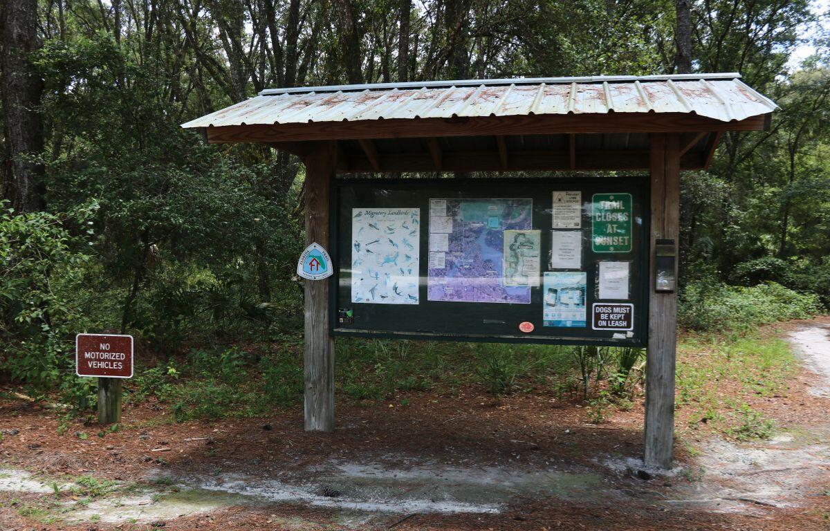 A wooden trailhead signboard with maps and notices stands at the start of a forest trail. Signs include "Trail Closes at Sunset" and "Dogs Must Be Kept on Leash." A smaller sign nearby says "No Motorized Vehicles." Trees and a dirt path surround the area.