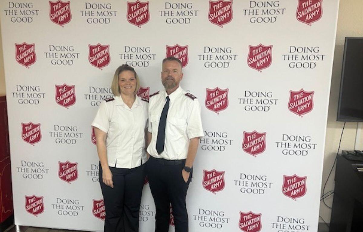 In the background a white screen with lettering and red shields with lettering, with a woman and man dressed in uniforms in the foreground.
