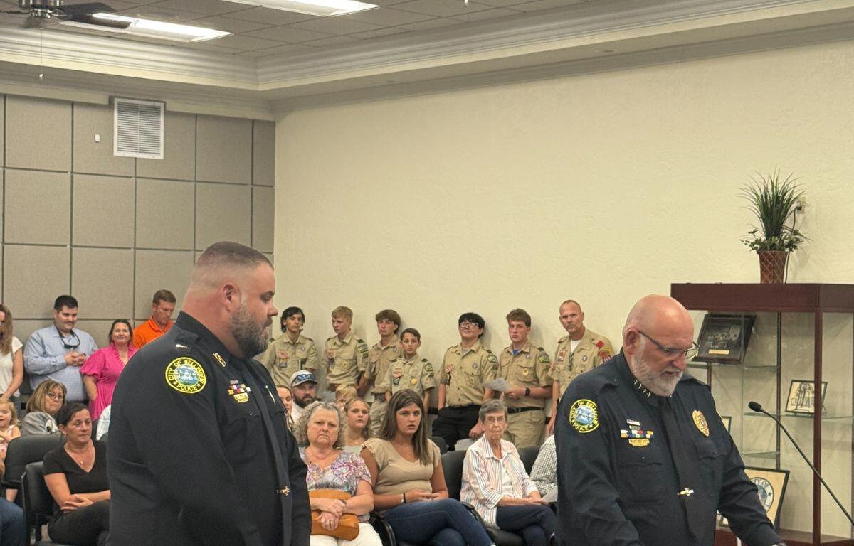A deep room. with people to the left, in the background and against the wall to the right, in the middle a man dressed in a law enforcement uniform, a man to his right also in a law enforcement uniform to his right at a podium with a microphone. In the foreground, there is the front of a dais.