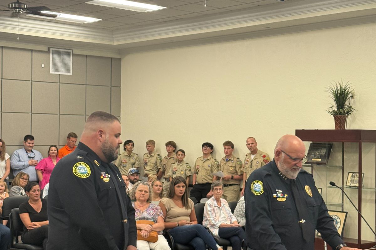 A deep room. with people to the left, in the background and against the wall to the right, in the middle a man dressed in a law enforcement uniform, a man to his right also in a law enforcement uniform to his right at a podium with a microphone. In the foreground, there is the front of a dais.