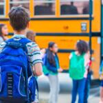 Children with backpacks stand near a yellow school bus on the first day of school.