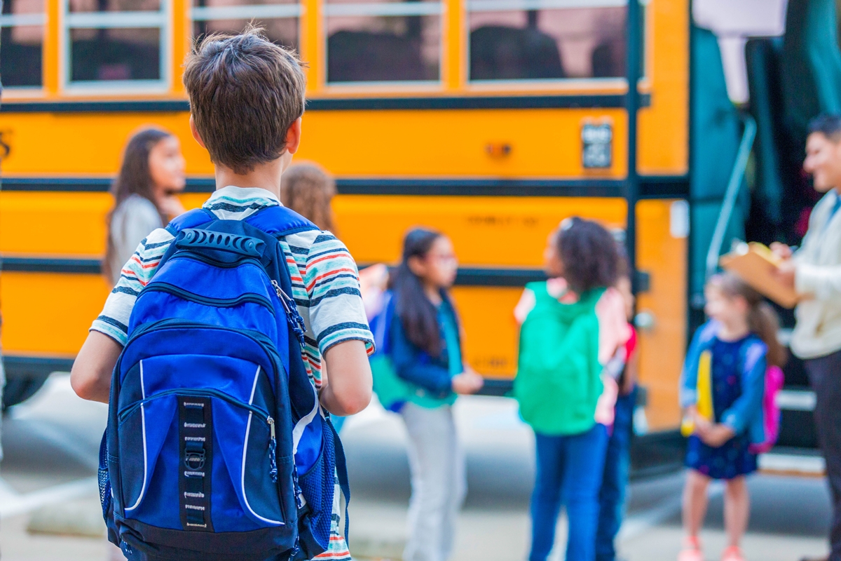 Children with backpacks stand near a yellow school bus on the first day of school.