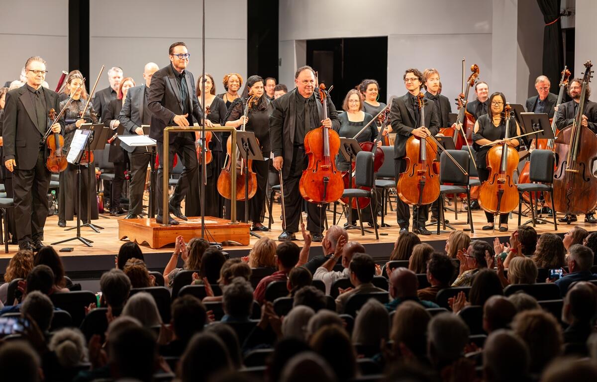 Matthew Wardell and members of the Ocala Symphony Orchestra stand on stage, smiling and holding instruments as the audience applauds.