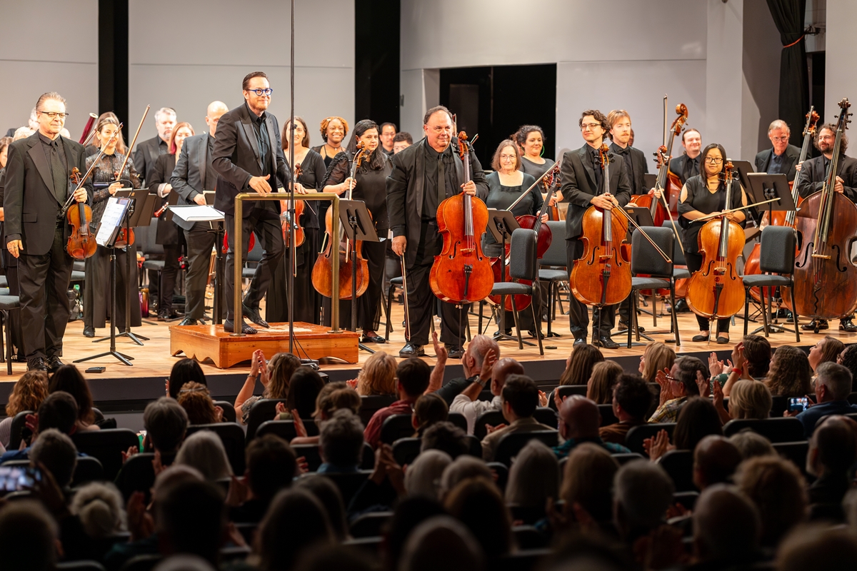 Matthew Wardell and members of the Ocala Symphony Orchestra stand on stage, smiling and holding instruments as the audience applauds.