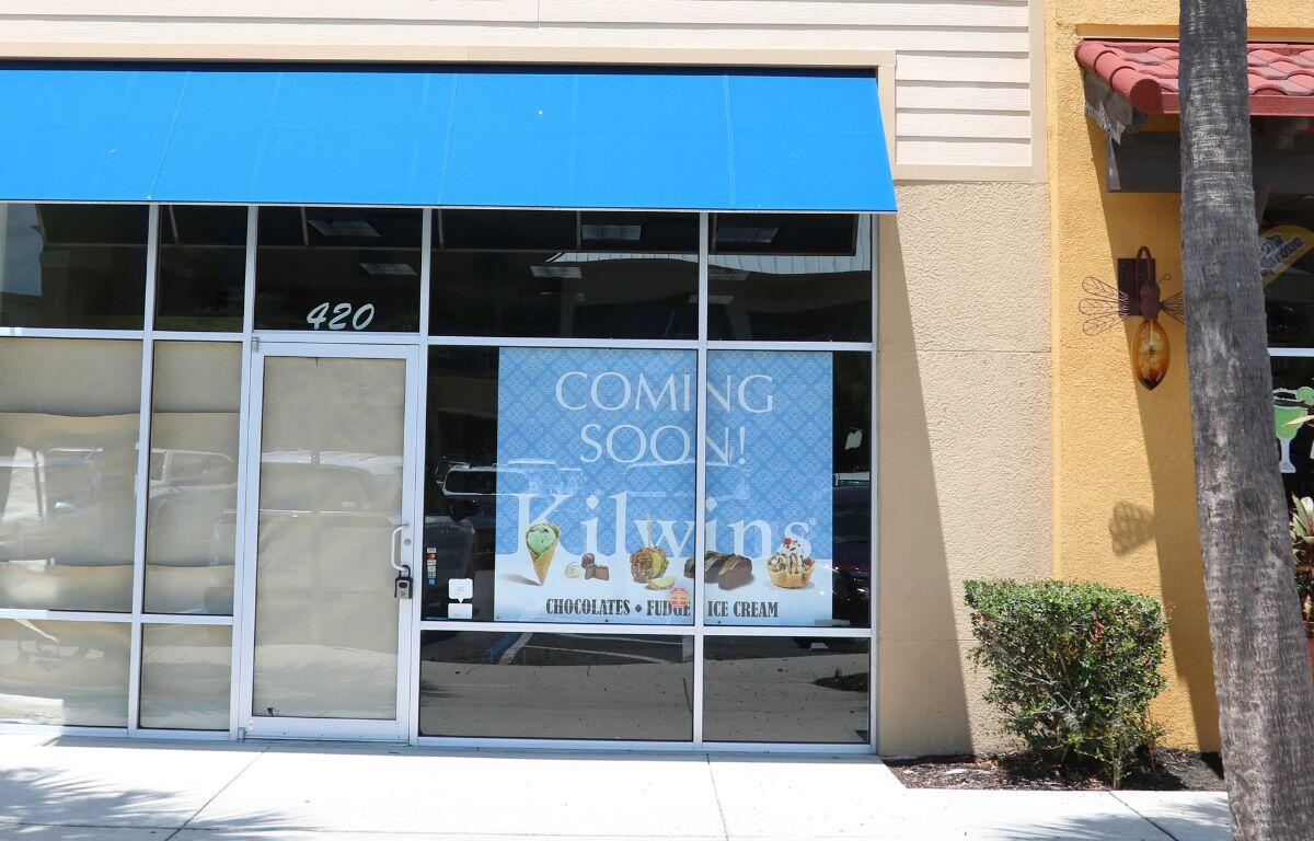 Storefront of an upcoming Kilwins location with a blue awning and large poster on the window reading “Coming Soon! Kilwins – Chocolates, Fudge, Ice Cream,” with images of sweet treats and the address number 420 above the door.