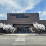 Front view of the in-progress Paddock Market at Paddock Mall in Ocala, featuring a modern dark brown exterior, large entrance doors, two white artificial trees, and two silver rearing horse sculptures on either side of the entrance.