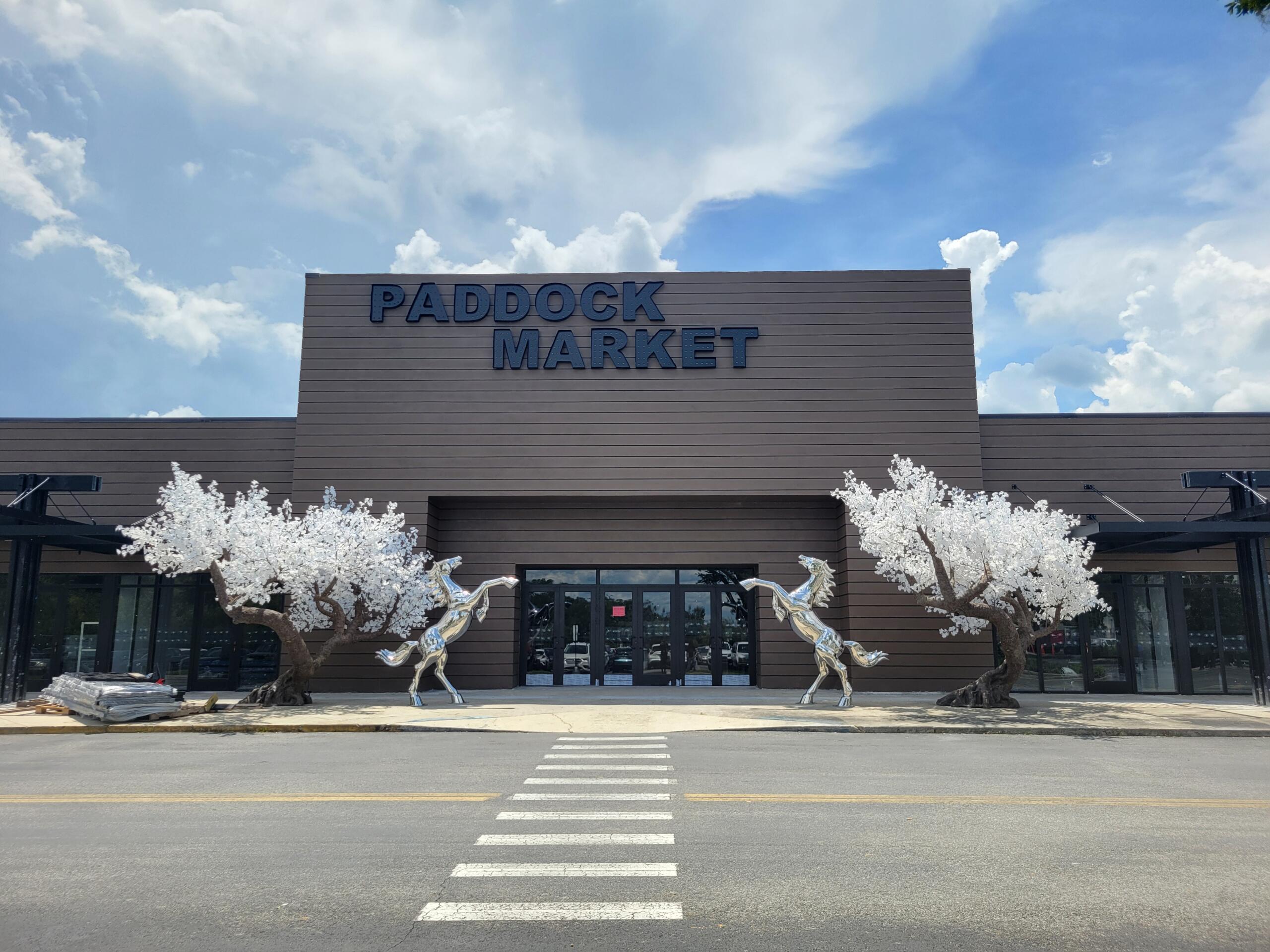 Front view of the in-progress Paddock Market at Paddock Mall in Ocala, featuring a modern dark brown exterior, large entrance doors, two white artificial trees, and two silver rearing horse sculptures on either side of the entrance.