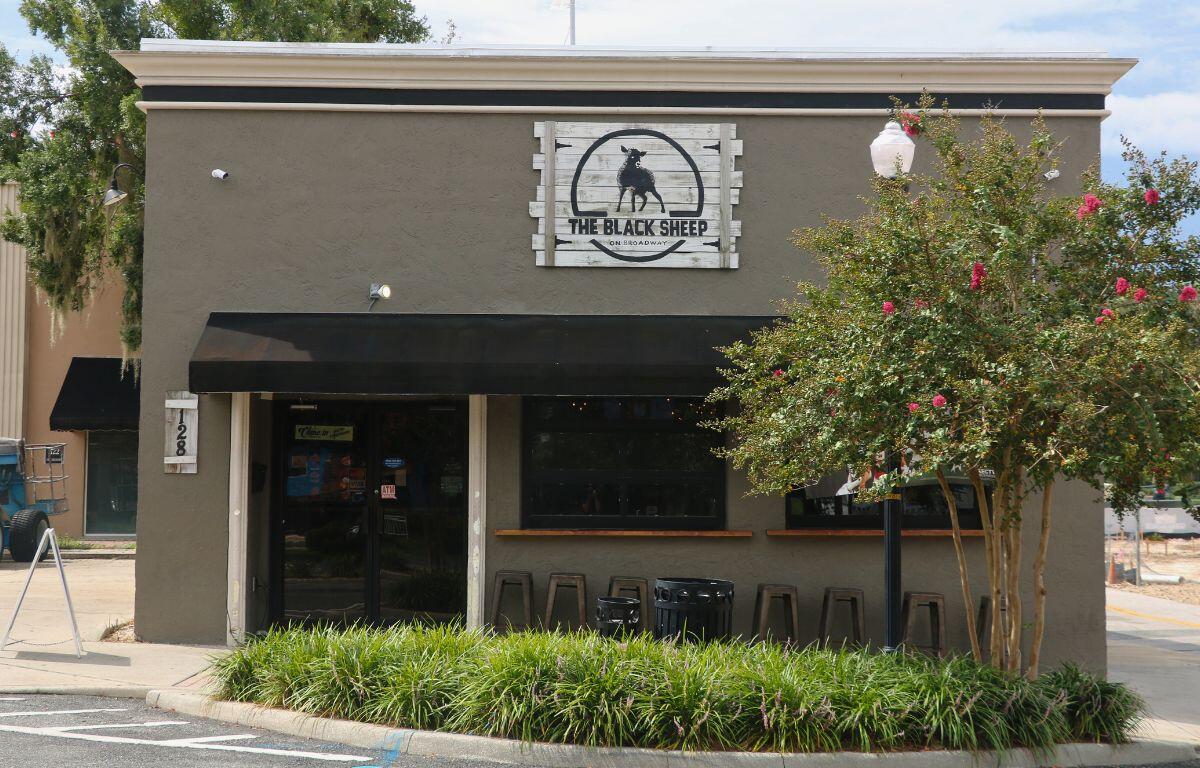 The exterior of a pub with grey, concrete walls sits at the corner of a street. The front of the building has a black awning above a door on the left and two windows to the right. Chair line the wall under the windows on the right. A sign toward the top of the building reads, "The Black Sheep" and a smaller sign to the right of the door reads "128." In front of the building is a planter with green plants filling the entirety of the planter with a tree toward the right and a light pole reaching up through the tree.