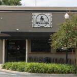 The exterior of a pub with grey, concrete walls sits at the corner of a street. The front of the building has a black awning above a door on the left and two windows to the right. Chair line the wall under the windows on the right. A sign toward the top of the building reads, "The Black Sheep" and a smaller sign to the right of the door reads "128." In front of the building is a planter with green plants filling the entirety of the planter with a tree toward the right and a light pole reaching up through the tree.
