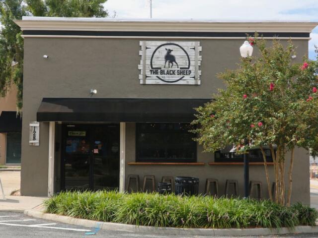 The exterior of a pub with grey, concrete walls sits at the corner of a street. The front of the building has a black awning above a door on the left and two windows to the right. Chair line the wall under the windows on the right. A sign toward the top of the building reads, "The Black Sheep" and a smaller sign to the right of the door reads "128." In front of the building is a planter with green plants filling the entirety of the planter with a tree toward the right and a light pole reaching up through the tree.