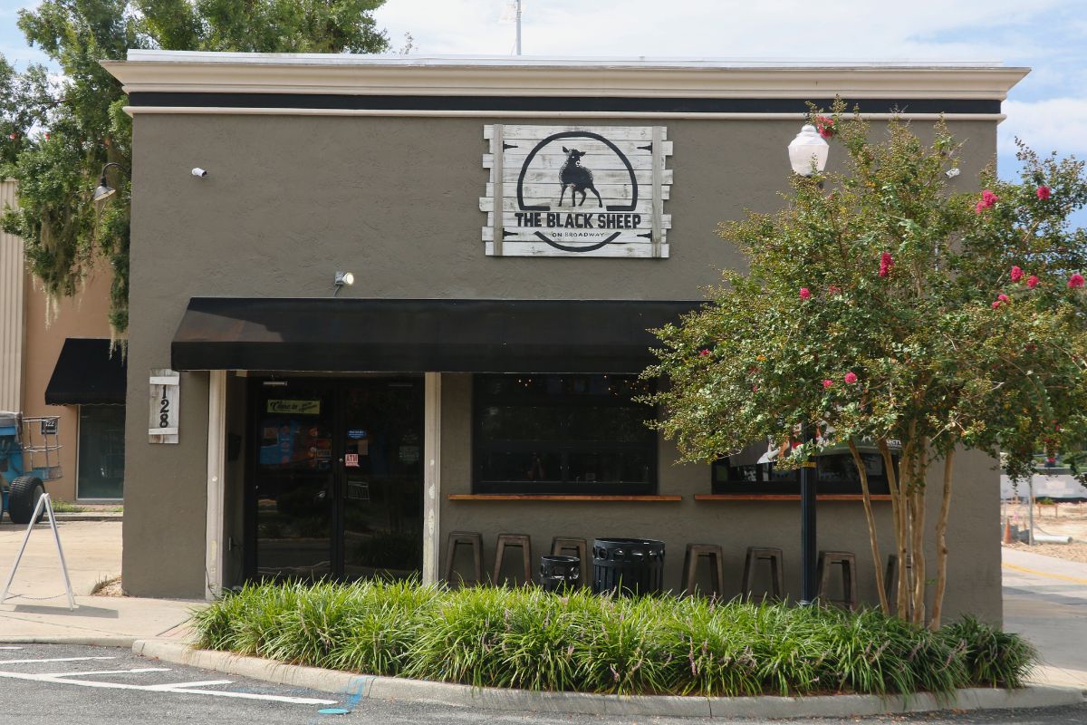 The exterior of a pub with grey, concrete walls sits at the corner of a street. The front of the building has a black awning above a door on the left and two windows to the right. Chair line the wall under the windows on the right. A sign toward the top of the building reads, "The Black Sheep" and a smaller sign to the right of the door reads "128." In front of the building is a planter with green plants filling the entirety of the planter with a tree toward the right and a light pole reaching up through the tree.