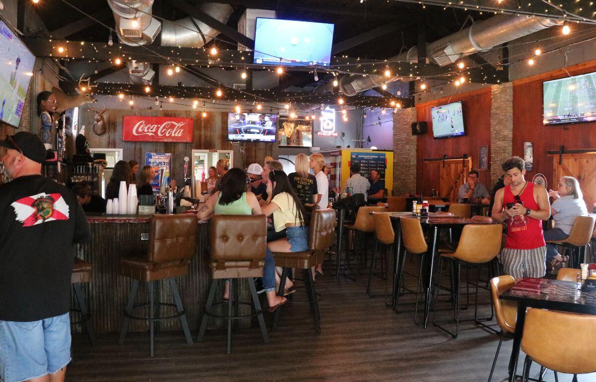 The inside of a pub has multiple televisions on the ceiling and walls. The walls are a mix of brick and wood. Guests sit at bar stool and tables, watching games on the televisions and mingling with each other.