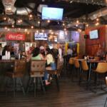 The inside of a pub has multiple televisions on the ceiling and walls. The walls are a mix of brick and wood. Guests sit at bar stool and tables, watching games on the televisions and mingling with each other.