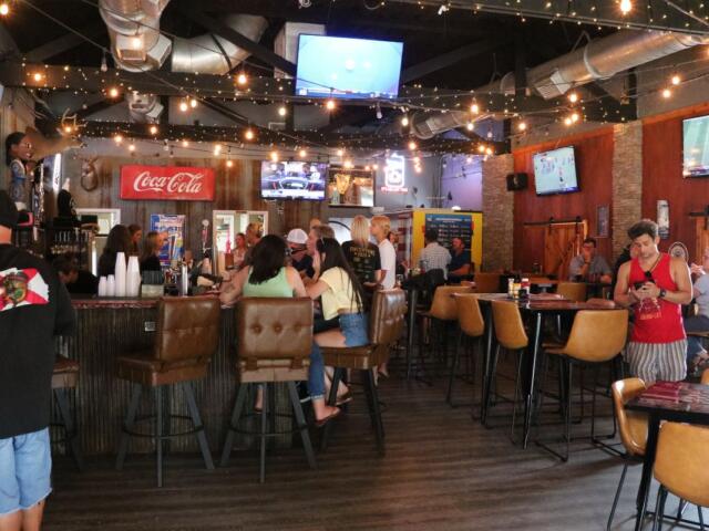 The inside of a pub has multiple televisions on the ceiling and walls. The walls are a mix of brick and wood. Guests sit at bar stool and tables, watching games on the televisions and mingling with each other.