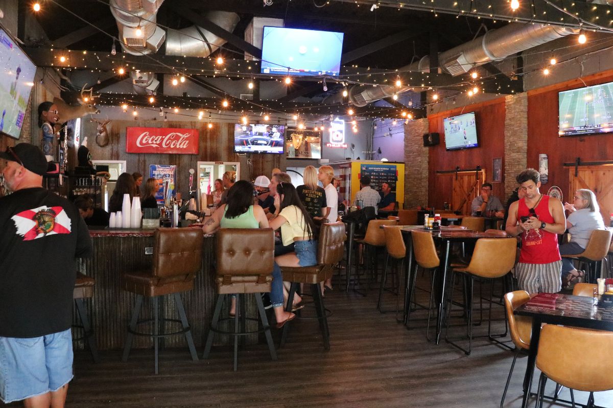 The inside of a pub has multiple televisions on the ceiling and walls. The walls are a mix of brick and wood. Guests sit at bar stool and tables, watching games on the televisions and mingling with each other.
