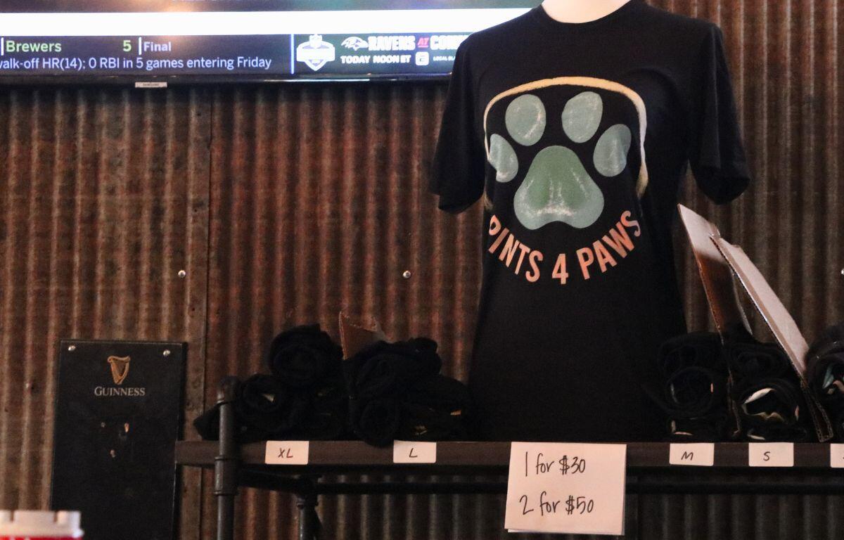 A shirt with a large, blue paw print reading, "Pints 4 Paws" with prices and sizes at the bottom sit atop a wood shelf.