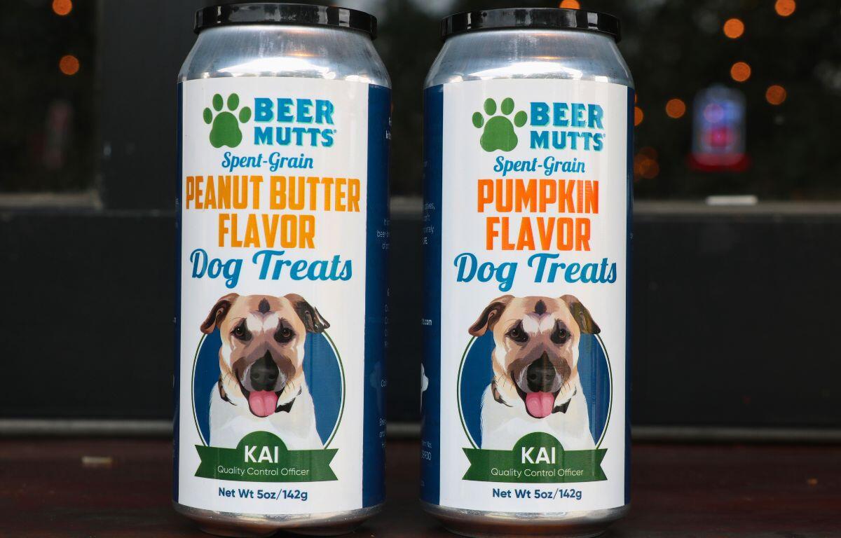 Two bottles of dog treats sit on a wood, pub counter. The container on the right reads, "Beer Mutts. Spent-Grain. Peanut Butter Flavor Dog Treats." The container on the right reads the same but with "Pumpkin Flavor" instead of "Peanut Butter Flavor."
