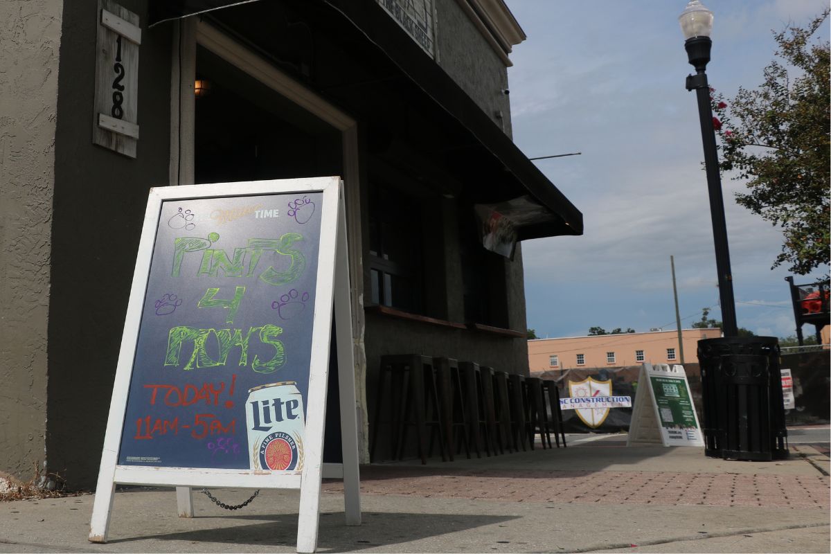 A sign outside of a grey, concrete pub on a street corner reads, "Pints 4 Paws," advertising a charity event at the pub in the background. A light pole, construction site, trash can and a tree stand in the background behind and in front of the building.