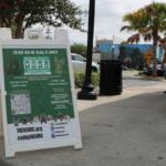 A white and green sign for the Humane Society of Marion County sits in front of a pub on the corner of a street with guests looking from a distance.