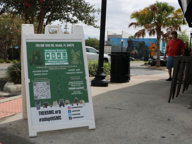 A white and green sign for the Humane Society of Marion County sits in front of a pub on the corner of a street with guests looking from a distance.
