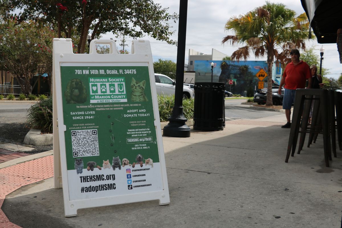 A white and green sign for the Humane Society of Marion County sits in front of a pub on the corner of a street with guests looking from a distance.