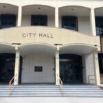 A building, with white trim, columns, a facade with lettering and steps leading toward the glass front doors.