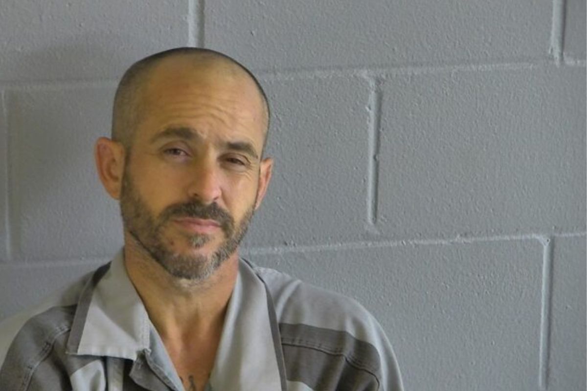 A man with a beard in a two-toned gray shirt of jail jump suit standing against the wall.
