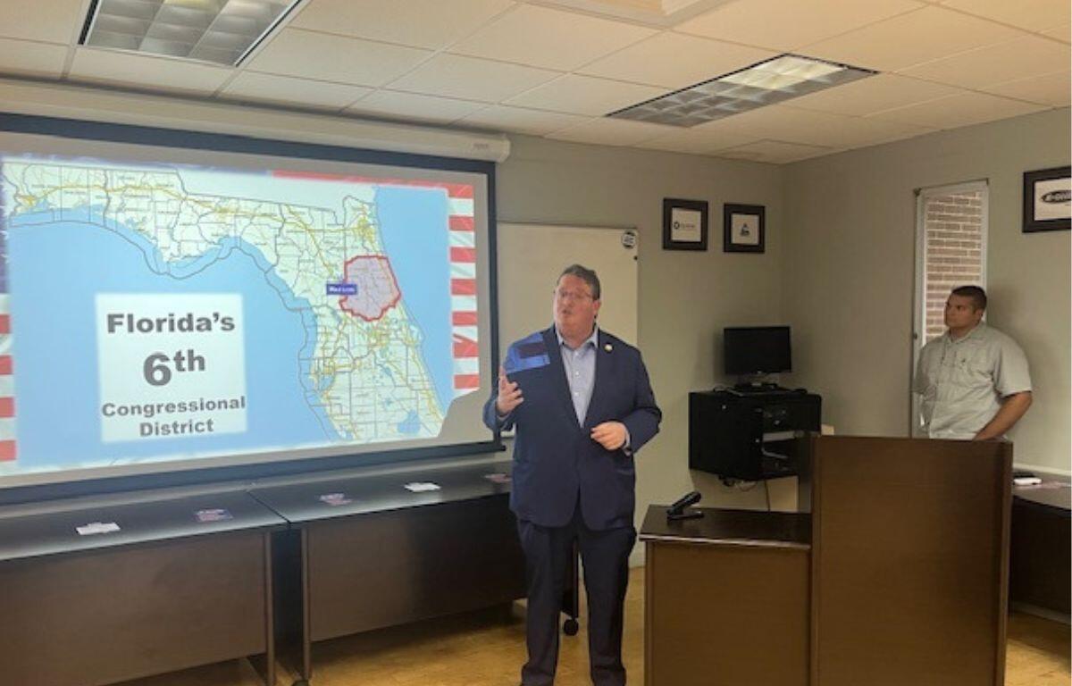A screen with a map on it, images, letters, graphics, with a ceiling overhead. A man in the middle of a room next to a podium. with a man standing on the far right.