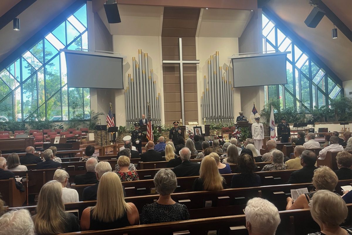 To the left, empty seats, two American flags, a video screen and organ pipes in the background, A cross in the middle and to the right more flags, another video screen and organ pipes. There are men behind the lecterns with microphones, on the left a man dressed in a suit, on the right a man dressed in a military uniform behind a lectern with a microphone. In front of them men in military uniforms, floral arrangements, a portrait of a man, and a table with items. In front of them in the foreground people seated in pews. 