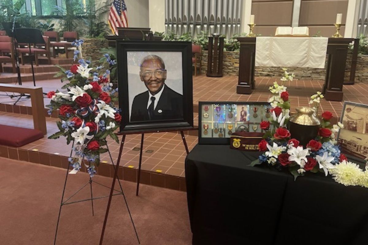 Floral display to the left of a portrait of a man in a suit and to the right in the foreground is a table with military decorations in shadow boxes, in the background is the American flag to the left, pipes to a pie organ, and to the right more pipes for a pie organ.