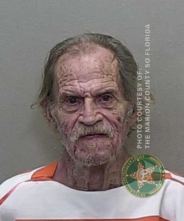 Mugshot of an older man with long gray hair and a thin mustache, wearing a white and orange prison uniform, against a gray background.