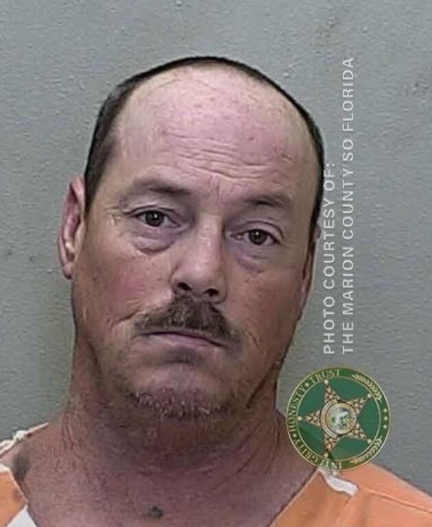 Mugshot of a man with short gray hair and facial hair, wearing a white and orange prison uniform, against a gray background.