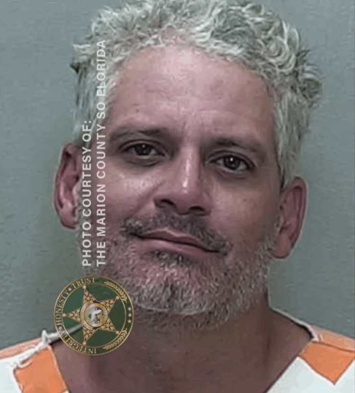 Mugshot of a man with short curly gray hair wearing an orange-and-white jail uniform, facing forward against a neutral background.