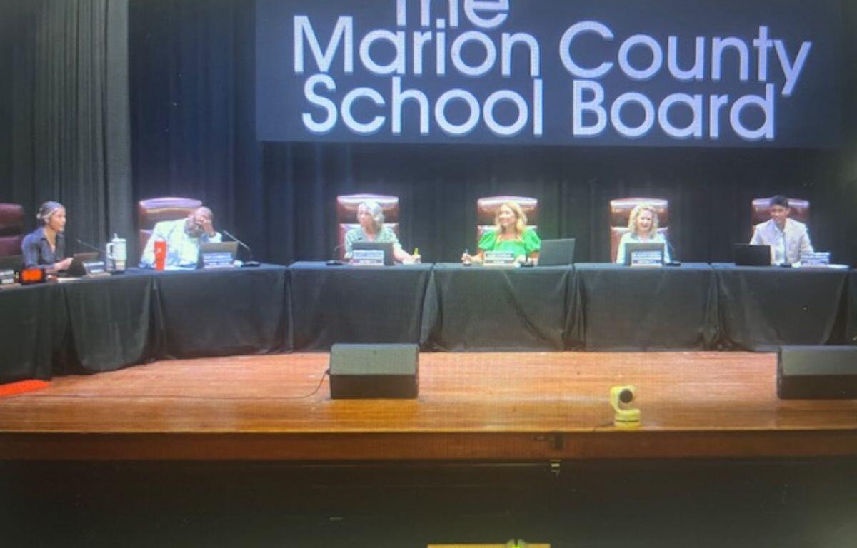 Well-dressed people sitting behind a dais, with a podiumin the foreground, with lighted lettering against a black fabric screen in the background.