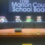 Well-dressed people sitting behind a dais, with a podiumin the foreground, with lighted lettering against a black fabric screen in the background.