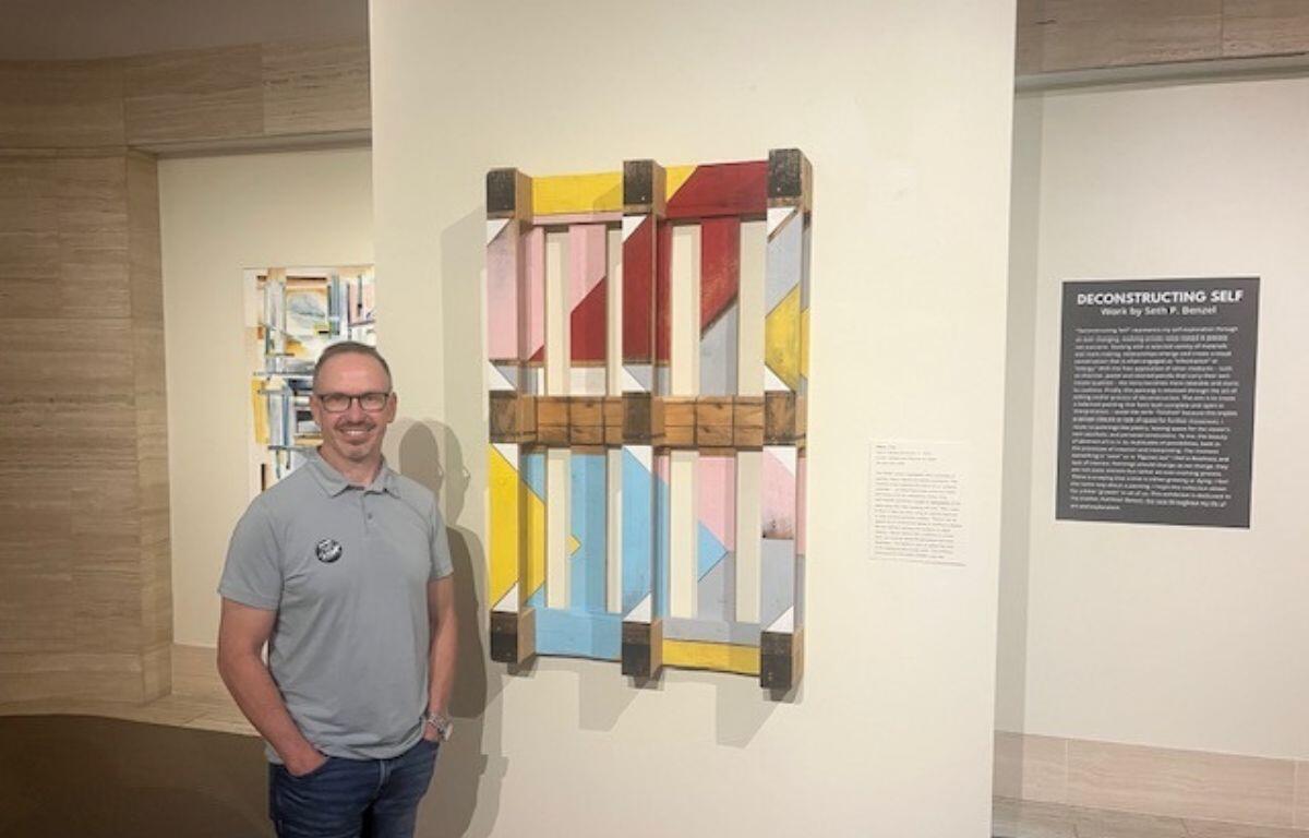 A man standing in front of a palette, that has been turned into a work of art, to his left, a painting, and to his right a placard with writing.