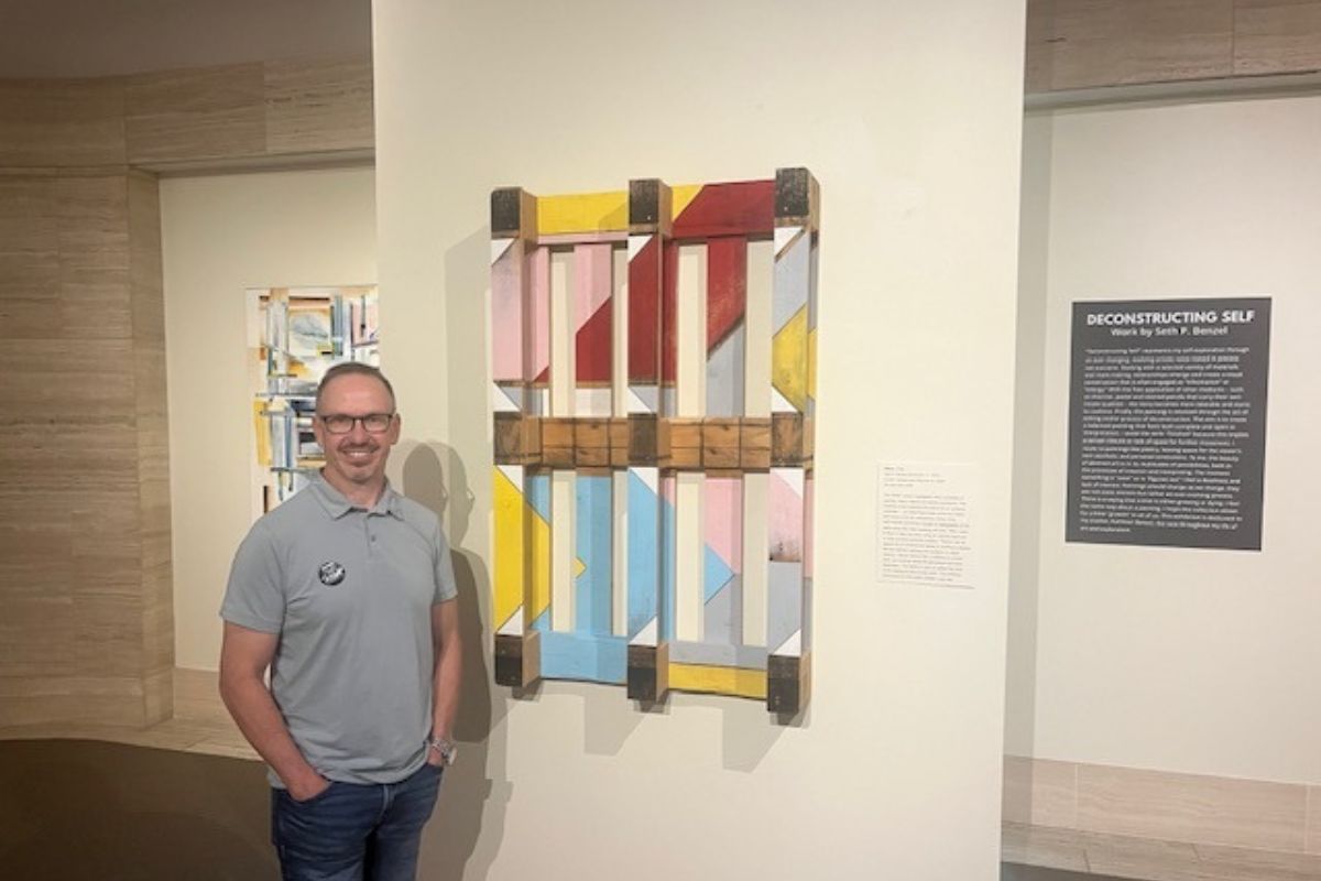 A man standing in front of a palette, that has been turned into a work of art, to his left, a painting, and to his right a placard with writing.
