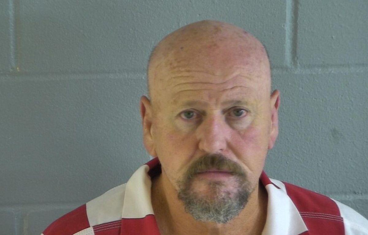 A bald man with a beard in an orange and white jump suit in front of a gray wall.